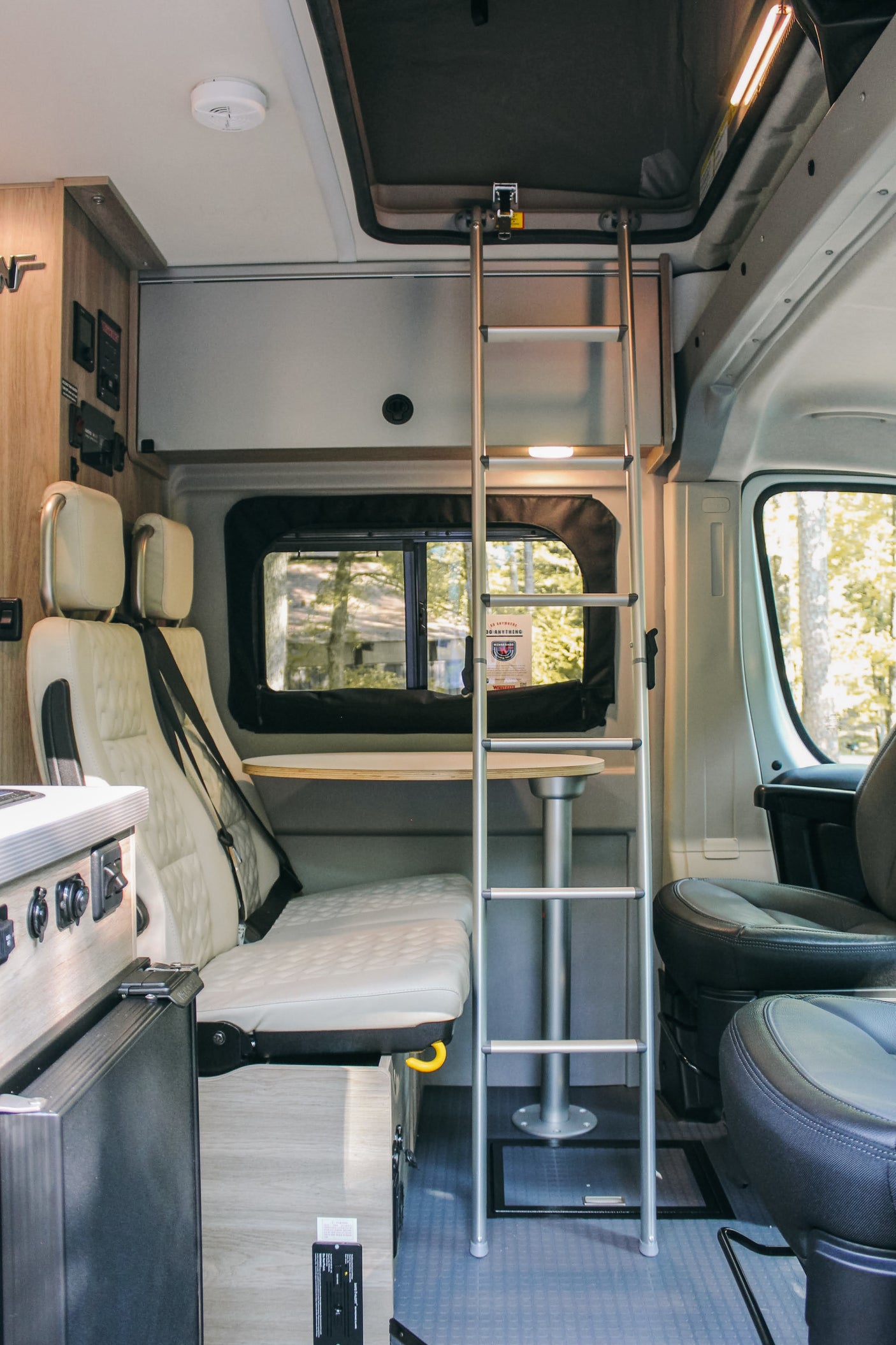 Inside this RV rental: cream dinette seating, a table, and a ladder to an overhead bunk. Front seats visible, ready for travel!