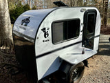 Enjoy nature with this white/black teardrop RV rental, a Koala by Rustic Trail, featuring an outdoor table in a sunny, wooded setting.