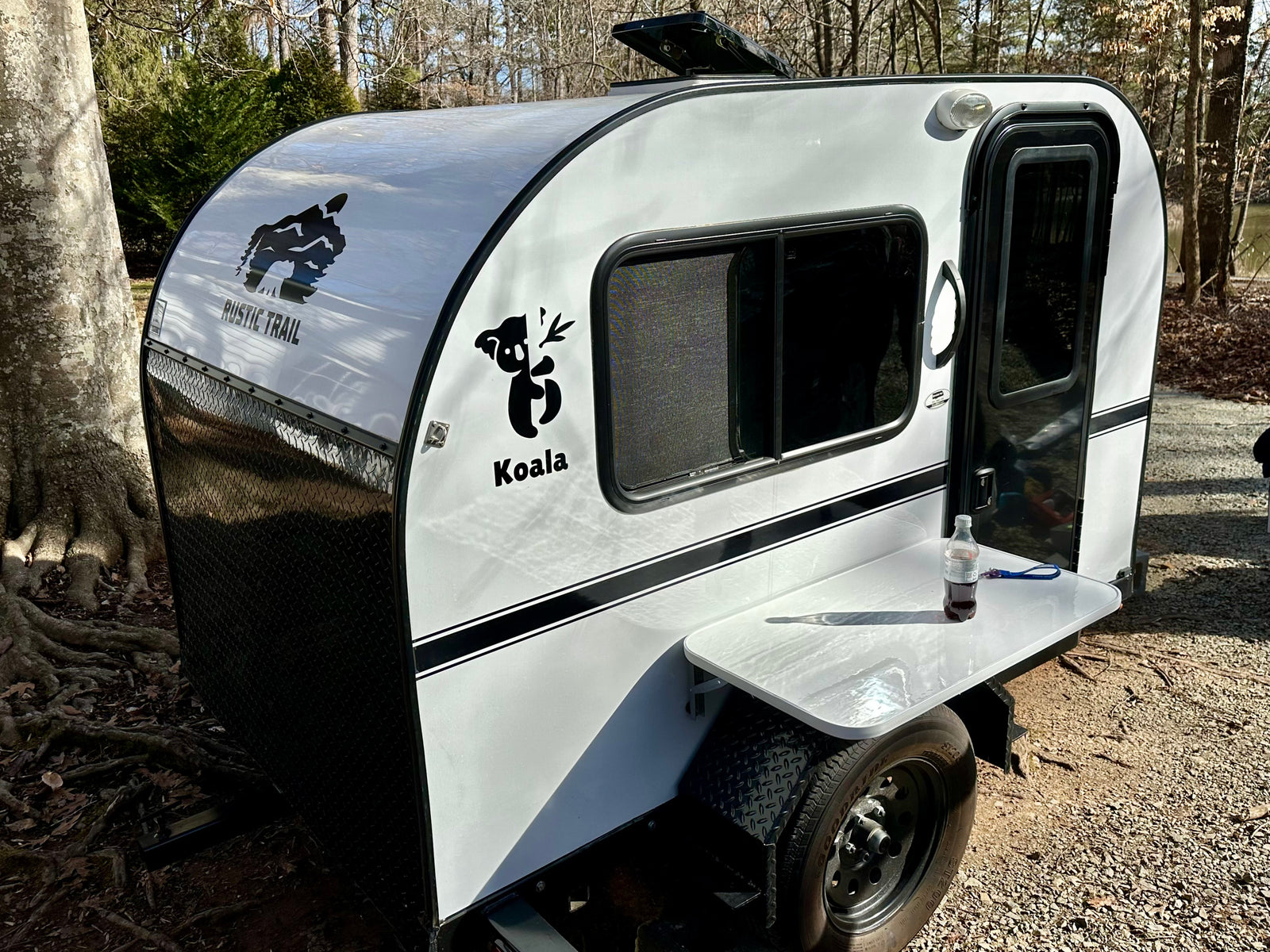 Enjoy nature with this white/black teardrop RV rental, a Koala by Rustic Trail, featuring an outdoor table in a sunny, wooded setting.