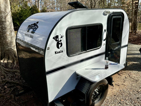 Enjoy nature with this white/black teardrop RV rental, a Koala by Rustic Trail, featuring an outdoor table in a sunny, wooded setting.
