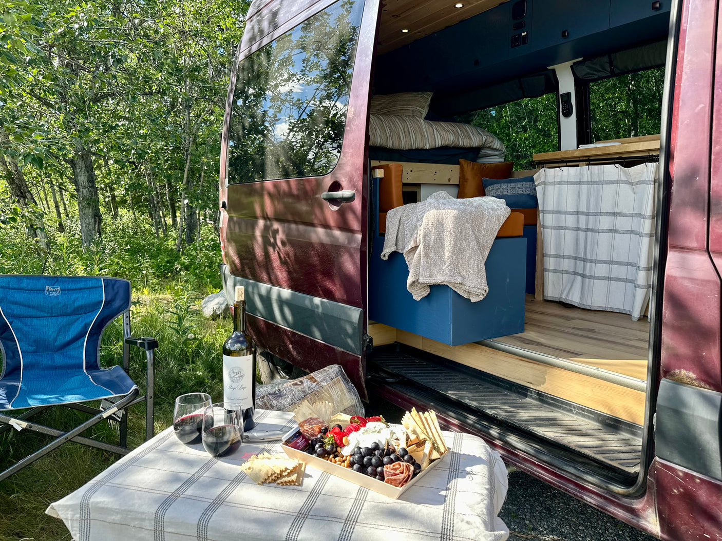 RV rental camper van parked in a sunny forest, with an outdoor charcuterie and wine setup beside it.