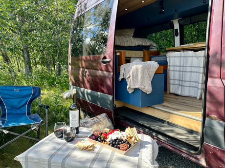 RV rental camper van parked in a sunny forest, with an outdoor charcuterie and wine setup beside it.