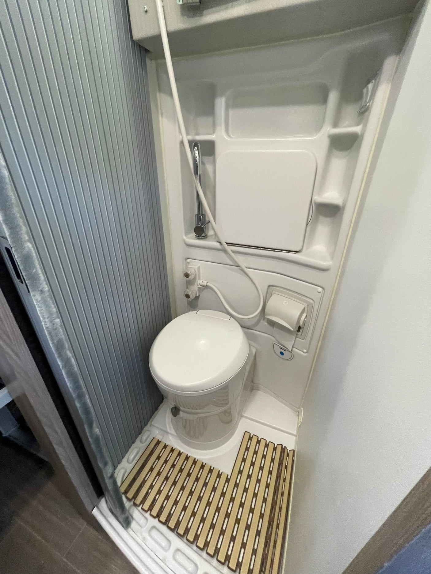 View of a compact RV rental bathroom: a white toilet, wall-mounted shower mixer, toilet paper dispenser, and a slatted mat on the floor.