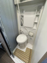 View of a compact RV rental bathroom: a white toilet, wall-mounted shower mixer, toilet paper dispenser, and a slatted mat on the floor.