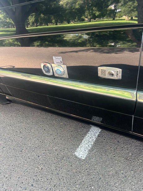 RV rental utility panel: power and fresh water hookups on a black RV, reflecting park trees and grass.