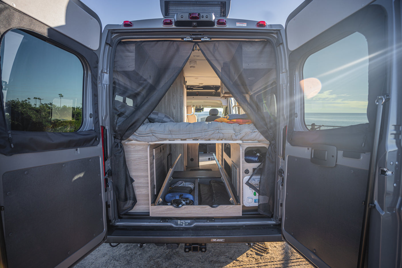 Rear view of an RV rental camper van, revealing its cozy interior with a bed, storage, and an ocean vista.
