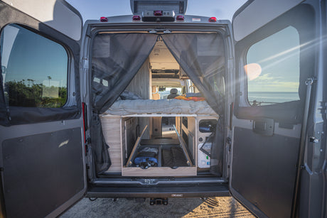 Rear view of an RV rental camper van, revealing its cozy interior with a bed, storage, and an ocean vista.