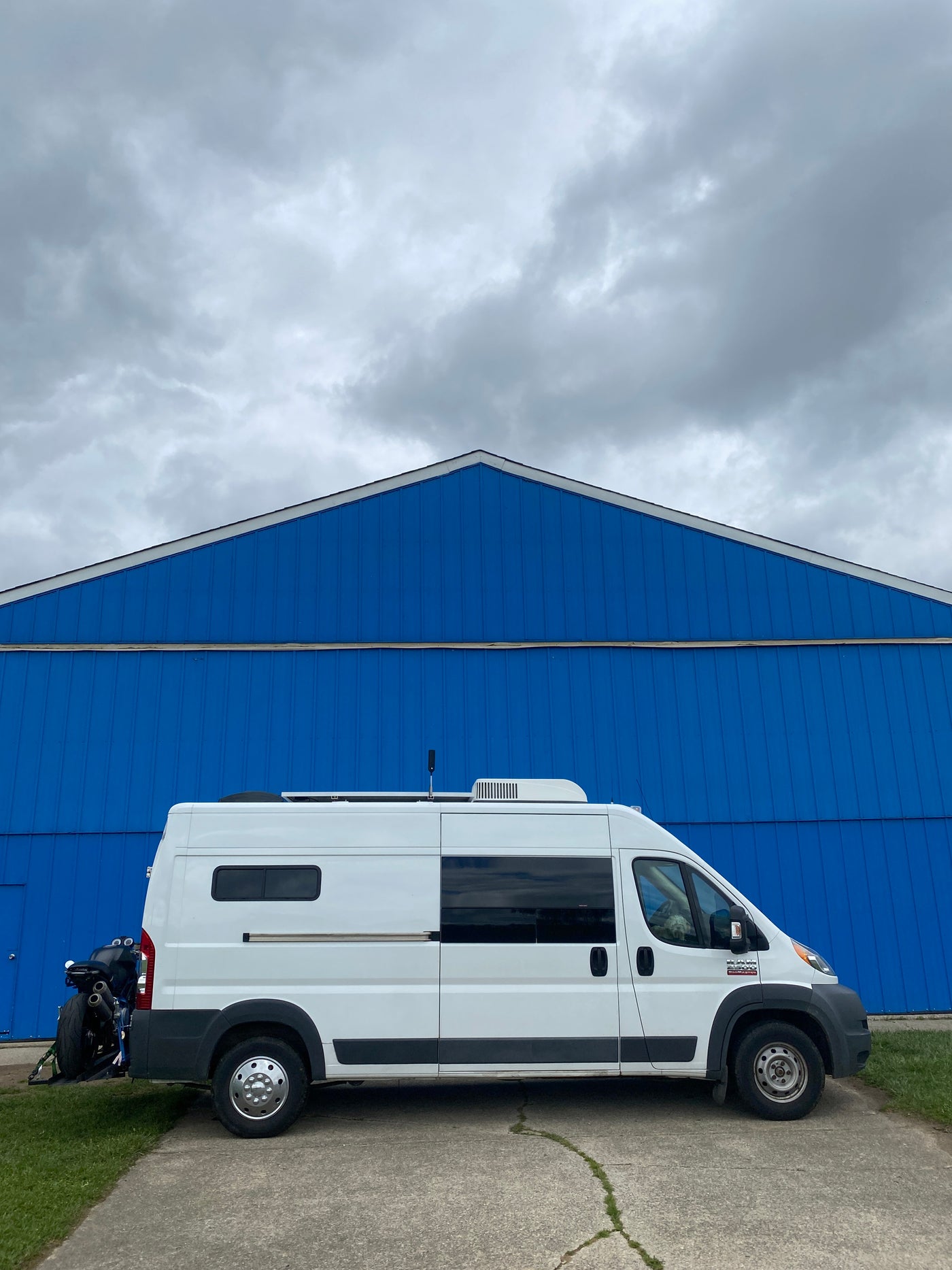 A white RV rental camper van with a rear-mounted motorcycle, parked by a vivid blue building under cloudy skies, ready for van life adventures. — photo 13