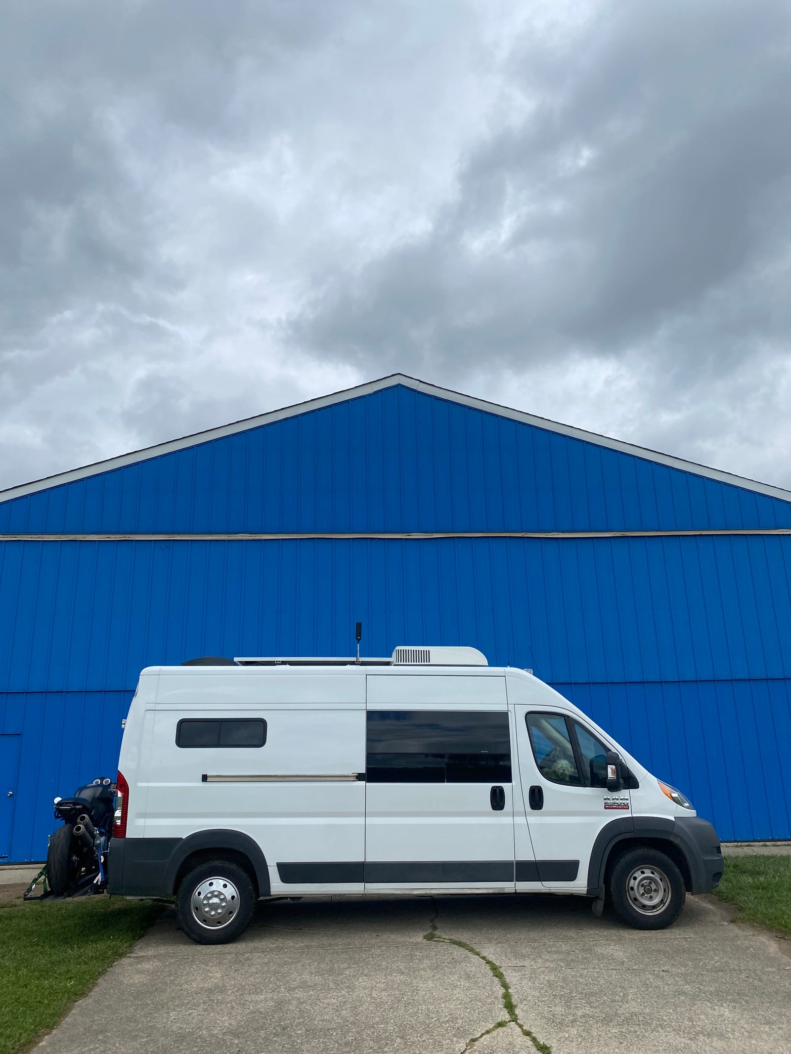 A white RV rental camper van with a rear-mounted motorcycle, parked by a vivid blue building under cloudy skies, ready for van life adventures.