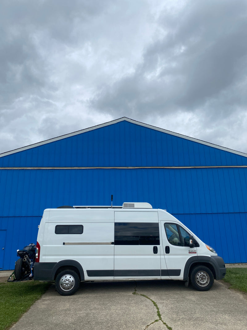 A white RV rental camper van with a rear-mounted motorcycle, parked by a vivid blue building under cloudy skies, ready for van life adventures. — photo 13