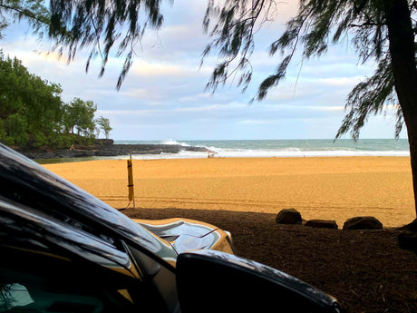 Coastal view from a parked RV rental: sandy beach, crashing waves, and lush trees under a cloudy sky.