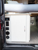 Inside an RV rental: A cozy bed, utility control panel with water/light/pump switches, power outlets, and a cabinet.
