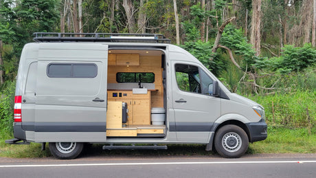 This light grey rv rental van, a custom Sprinter, reveals a warm wood interior with kitchen and toilet, parked roadside.