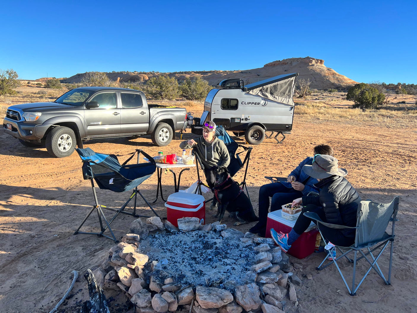 Friends and a dog gathered around a fire pit at a desert camp with an rv rental and pickup truck.