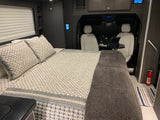 Inside a modern RV rental: a patterned bed with a dark blanket, front white seats with a laptop table, and safety equipment.