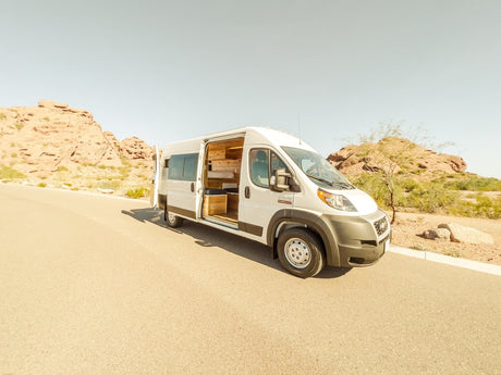 White camper van RV rental, side door open to wood interior, parked on a desert road with rocky hills.