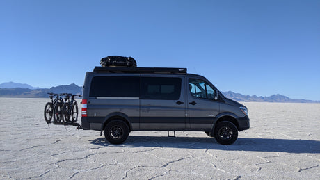 Explore with an RV rental! Gray van with bikes on vast white salt flats, mountains in distance, clear blue sky.