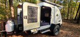 This rv rental is a cream teardrop trailer at a wooded campsite, showing its open door, interior bed, and AC unit.