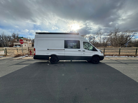 White camper van, resembling an RV rental, parked on a road under a sunlit, cloudy sky.