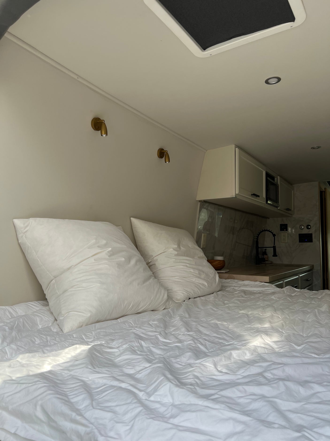 Comfortable RV rental interior: a bed with white linens, brass reading lights, and a compact kitchen area.