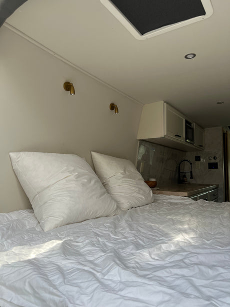 Comfortable RV rental interior: a bed with white linens, brass reading lights, and a compact kitchen area.