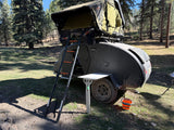 An RV rental teardrop camper with a rooftop tent, set up in a sunny pine forest for camping and outdoor adventures.