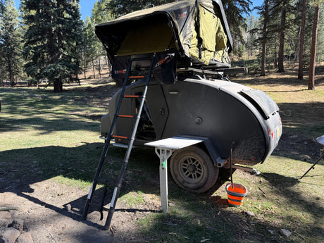 An RV rental teardrop camper with a rooftop tent, set up in a sunny pine forest for camping and outdoor adventures.