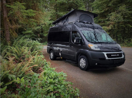 Dark grey camper van with pop-top extended, parked by lush forest. Your ideal rv rental for nature adventures.