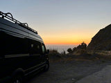 Dark RV rental van parked on a coastal pull-off, silhouetted against a vibrant sunset over the ocean and distant hills.