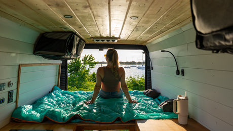 A woman sits on a turquoise sleeping bag in a cozy RV rental van, looking out at boats on the water.