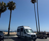 White camper van (potential RV rental) with door open, parked by a sunny beach with palm trees and beachgoers.