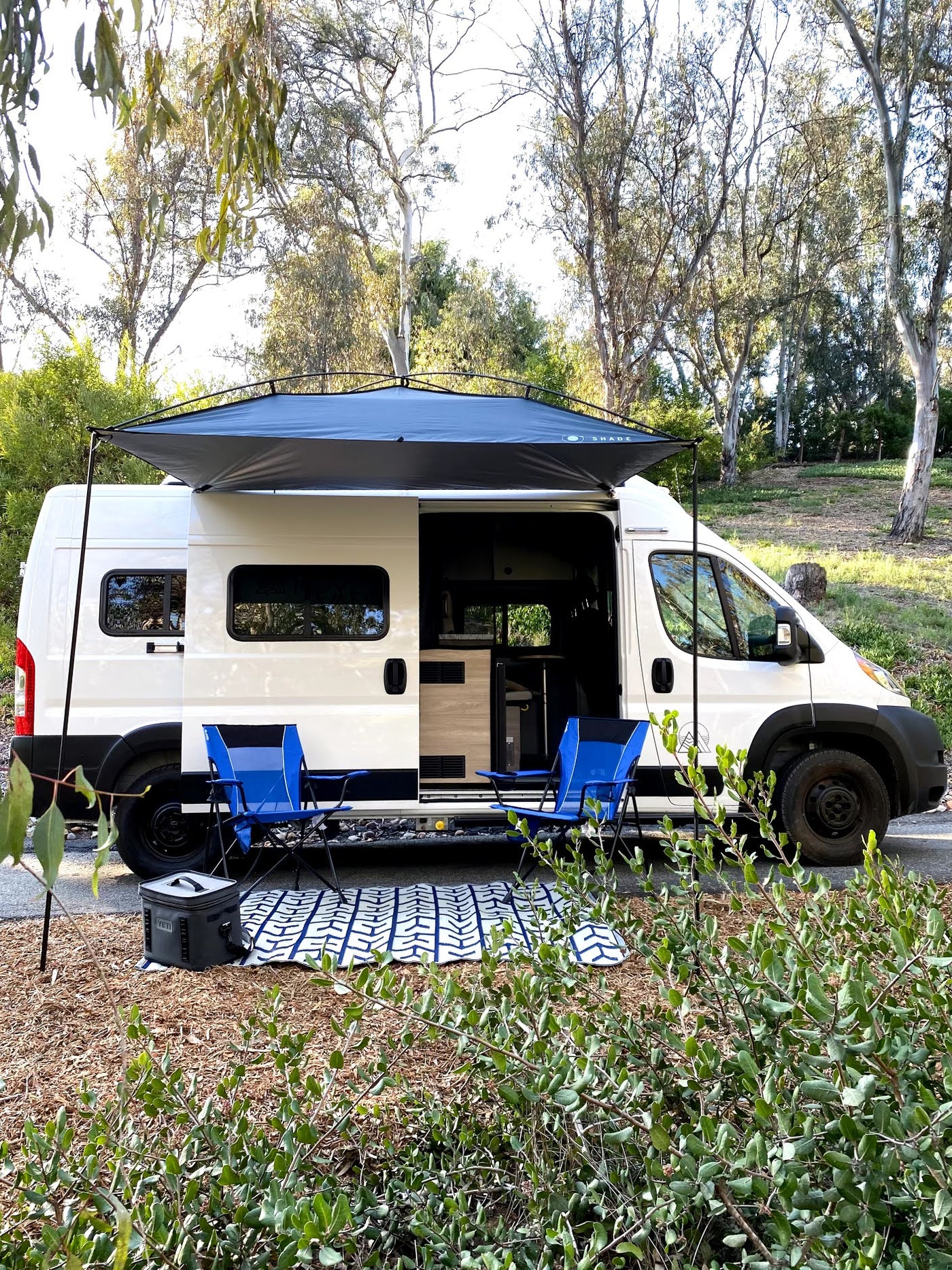 Scenic rv rental setup: White camper van with awning, blue chairs, and cooler, ready for outdoor comfort in nature. — photo 2