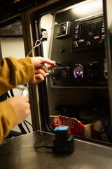 Inside an RV rental: a hand plugs a device into USB ports on a control panel, showing Webasto heater & power gauge. Cereal box nearby.