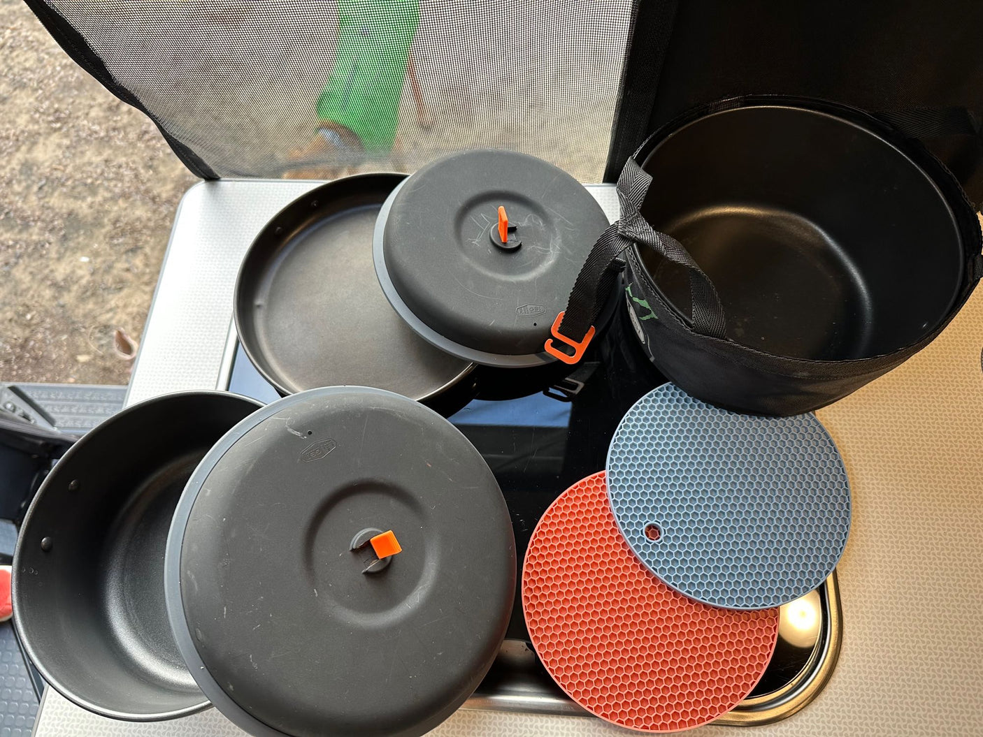 RV rental ready: Compact camping cookware, with black pots, pans, and lids with orange handles. Plus, blue & red silicone trivets.