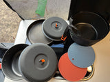 RV rental ready: Compact camping cookware, with black pots, pans, and lids with orange handles. Plus, blue & red silicone trivets.