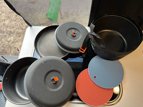 RV rental ready: Compact camping cookware, with black pots, pans, and lids with orange handles. Plus, blue & red silicone trivets.