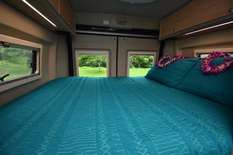 This inviting RV rental interior features a cozy bed with a teal quilt, floral leis, and expansive nature views.