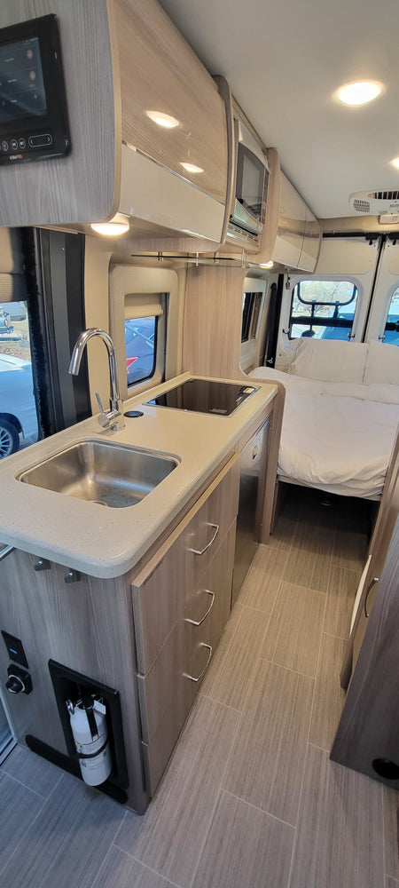 Modern RV rental interior: Compact kitchen with sink, cooktop, overhead storage, and a cozy bed area in back. Great for adventures!