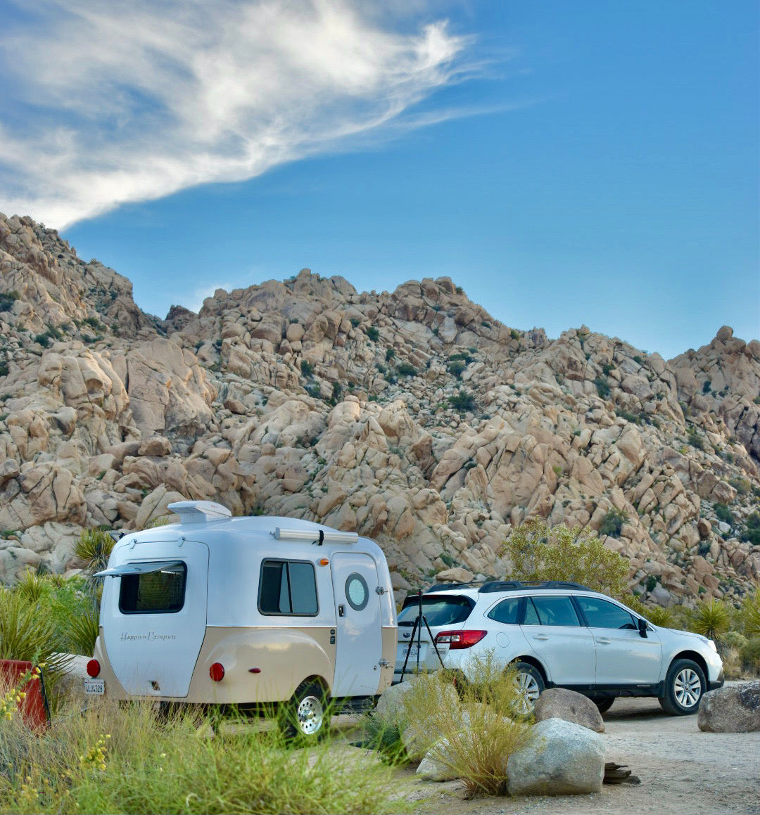 An RV rental Happier Camper travel trailer and Subaru Outback are parked in a rocky desert campsite.