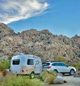 An RV rental Happier Camper travel trailer and Subaru Outback are parked in a rocky desert campsite.