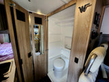 Interior of an rv rental, showing a compact bathroom with toilet and shower stall. A bed and small sink are to the left.