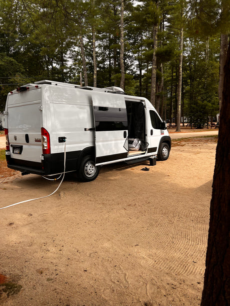 White RV rental van parked on a sandy campsite amidst tall pines. Side door open, utility hookup visible.