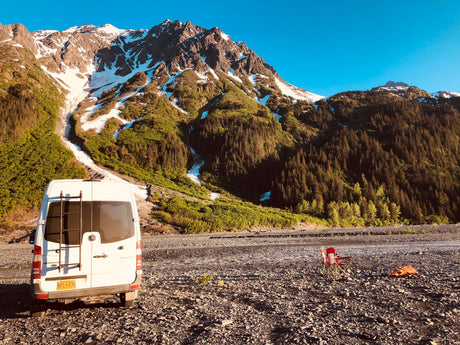 This RV rental camper van rests on a rocky Alaskan riverbed, backed by snow-capped mountains, next to a campfire.