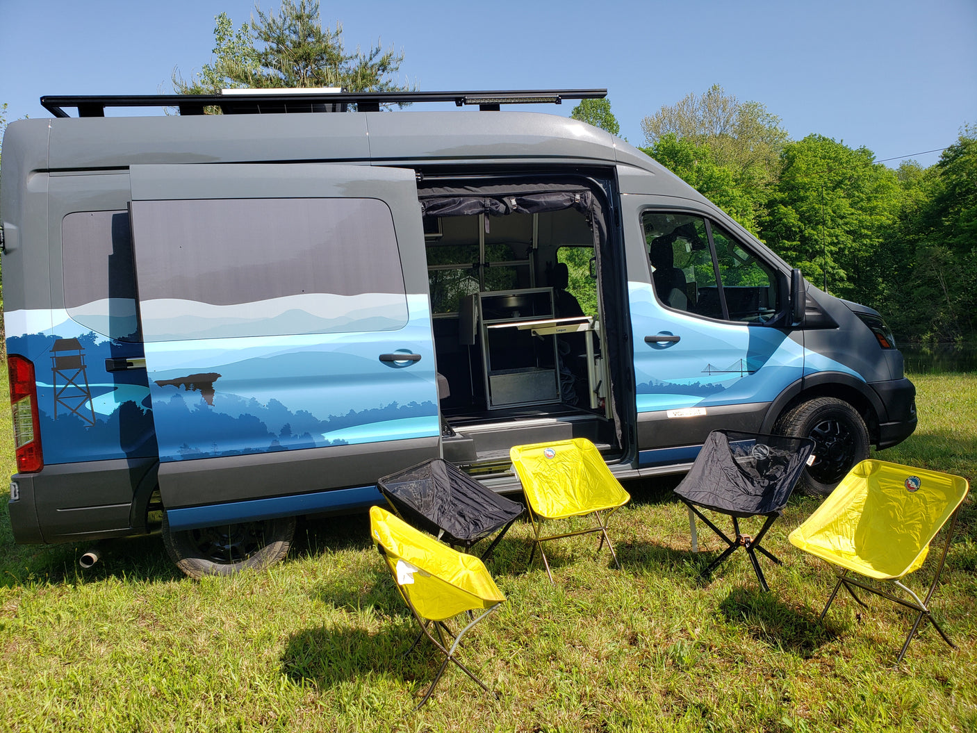 Custom rv rental van with scenic blue graphics and open door, parked on grass with camping chairs. Ideal for adventures.