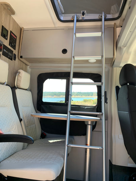Scenic lake view from a comfortable RV rental interior with dinette seating, control panels, and a ladder to an upper sleeping area.