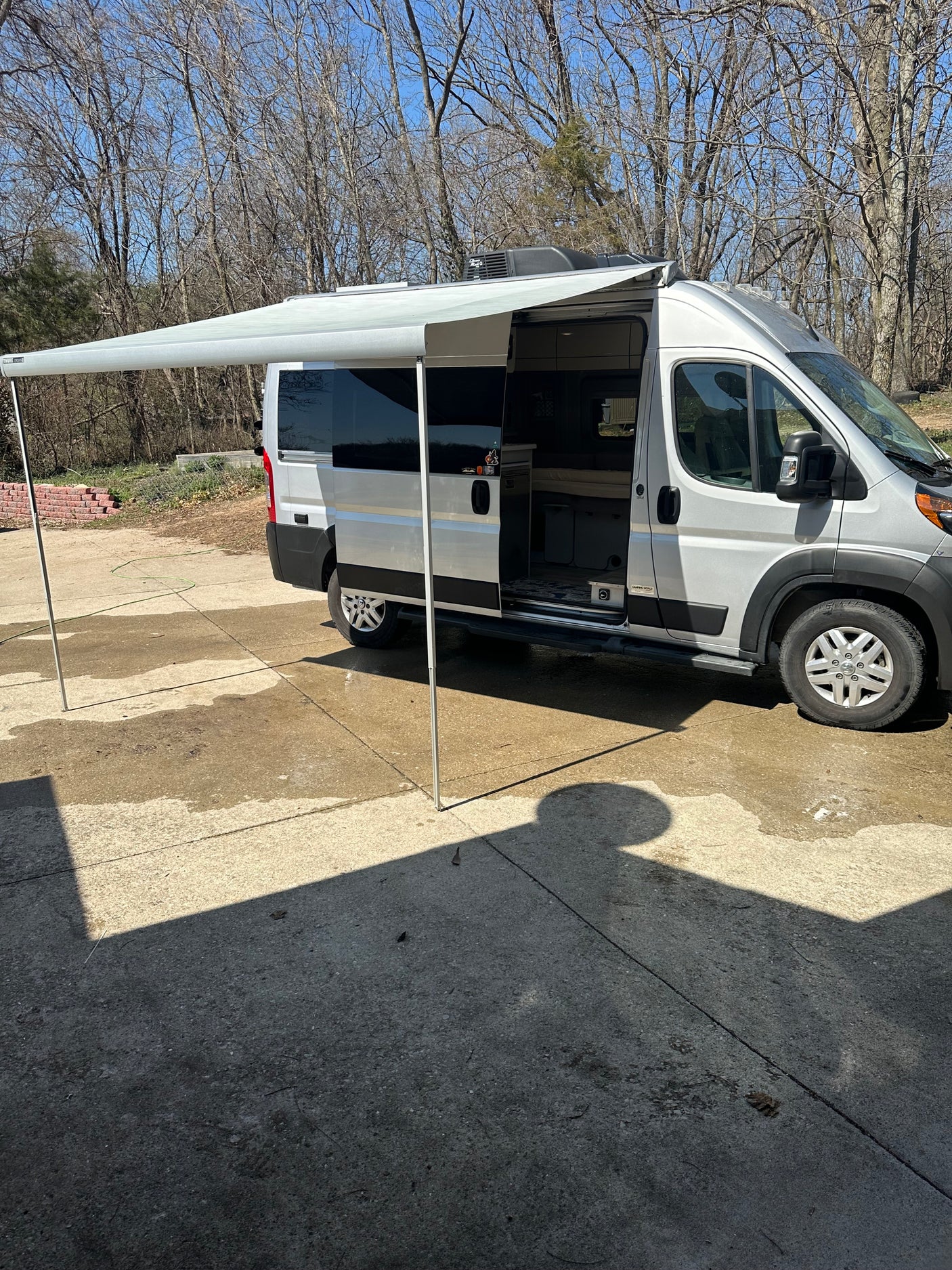 A silver camper van (RV rental) with its awning extended, parked on concrete on a sunny day with bare trees.