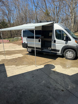 A silver camper van (RV rental) with its awning extended, parked on concrete on a sunny day with bare trees.