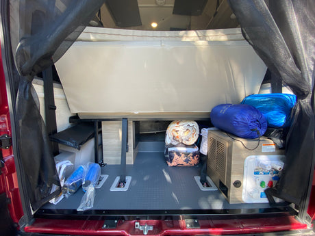 RV rental camper van interior: rear storage with folded bed, bedding, gear, utility panel, and dark mesh screens.