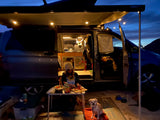 Evening at the RV rental: A person preps food outside their camper van, dog nearby, under a lighted awning.