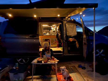 Evening at the RV rental: A person preps food outside their camper van, dog nearby, under a lighted awning.